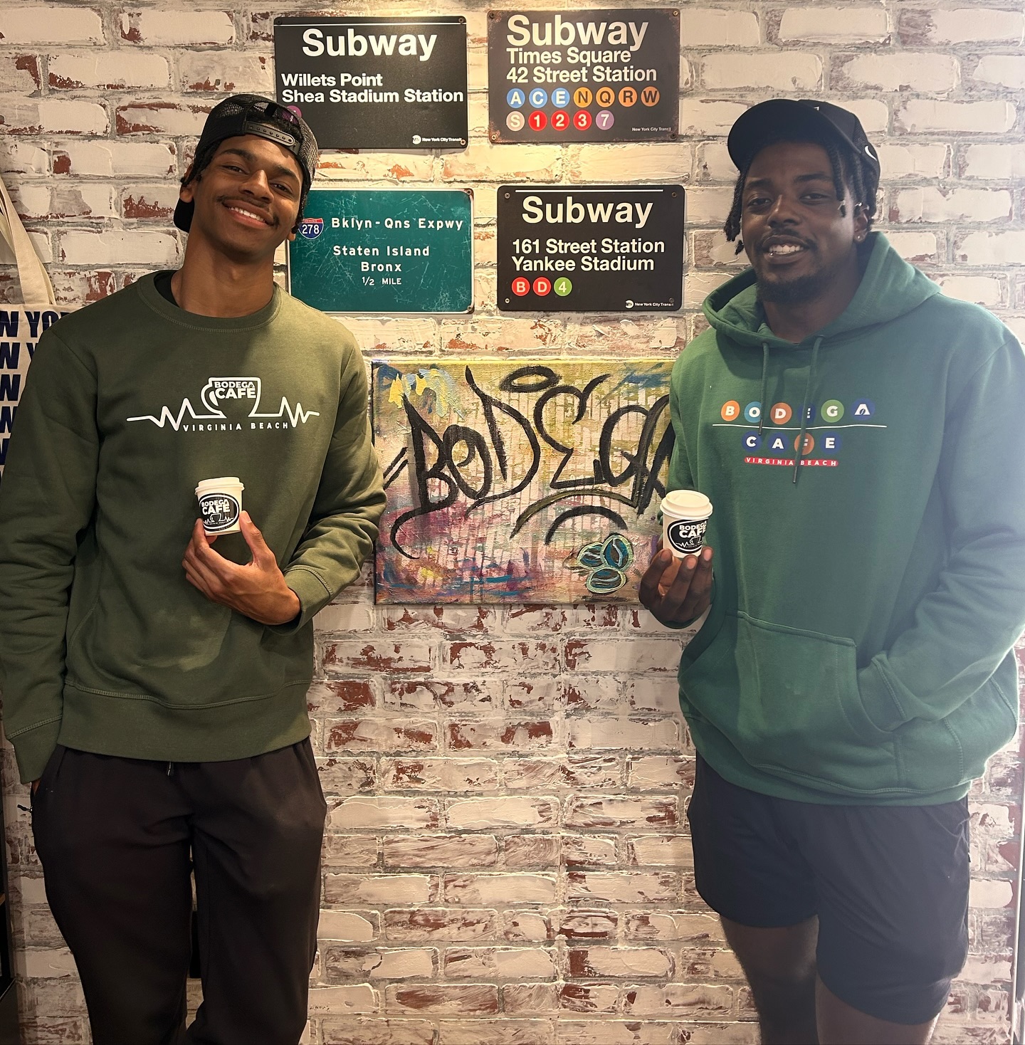 Two staff in Bodega Café hoodies in front of NYC subway wall art